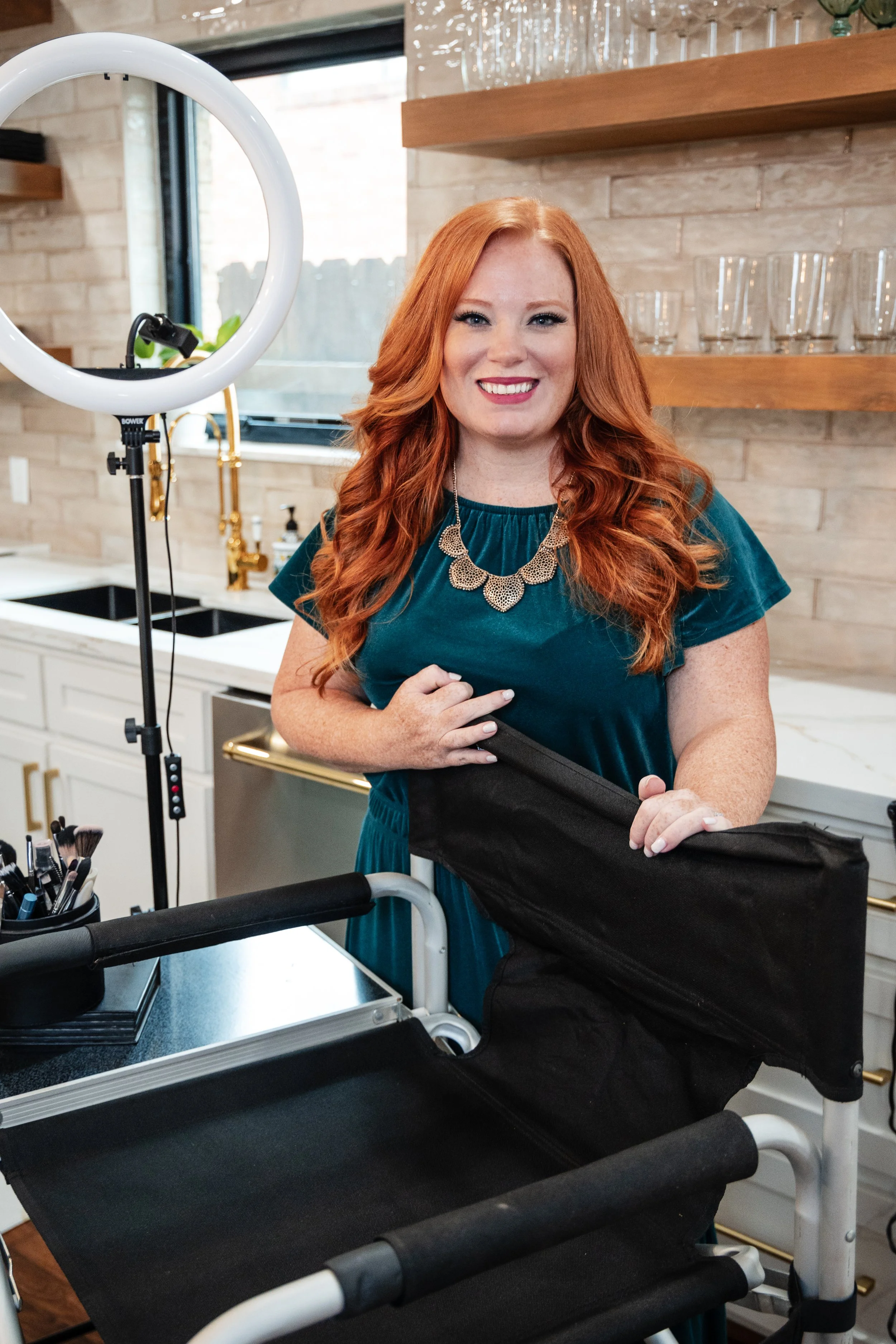 A woman with long, curly red hair, wearing a teal dress and a statement necklace, smiling in a modern kitchen with a makeup cart, ring light, and shelves with glasses.