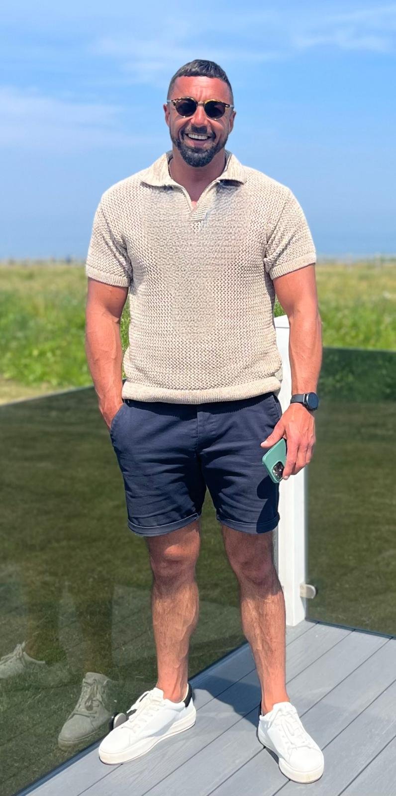 A man smiling outdoors on a sunny day, wearing sunglasses, a beige short-sleeve shirt, navy shorts, white sneakers, and a smartwatch, holding a phone in his right hand.