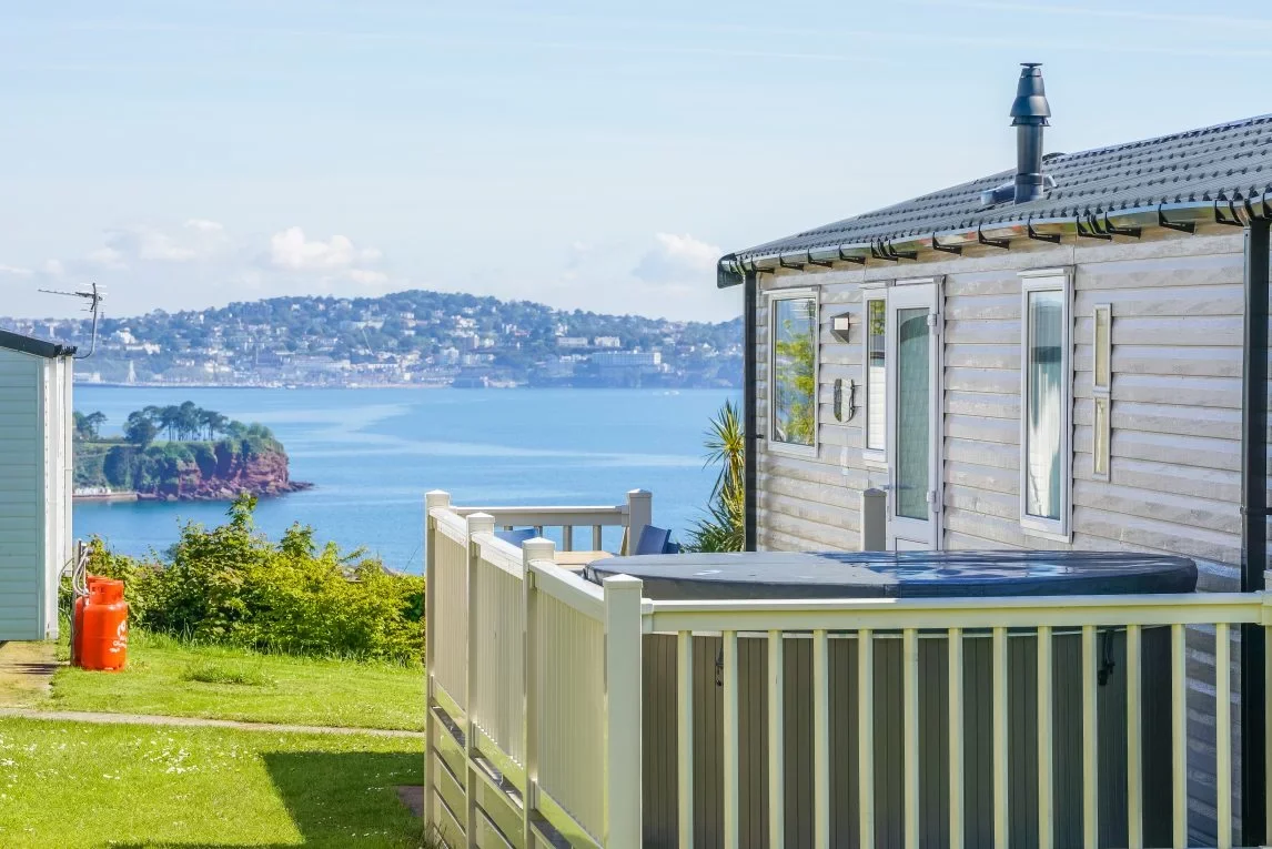 View of a house with a backyard overlooking a bay or river, with a dock, trees, and a distant cityscape on the hillside.