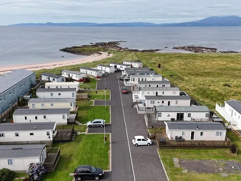 A coastal residential community with houses and parked cars near the water, overlooking a calm sea and distant mountains.