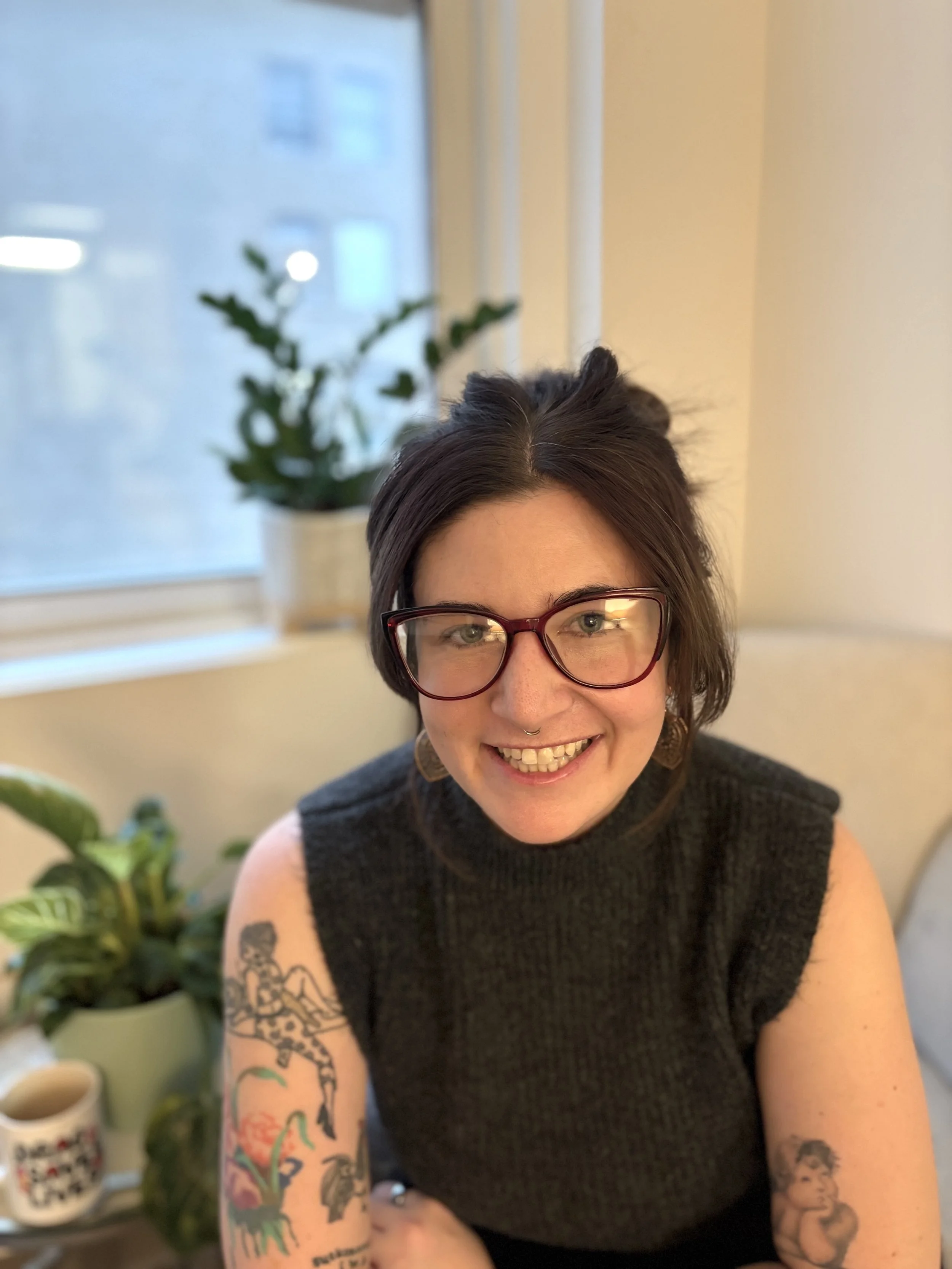 A woman with short dark hair, glasses, and tattoos on her arms smiling at the camera in an indoor setting with a window and potted plants behind her.