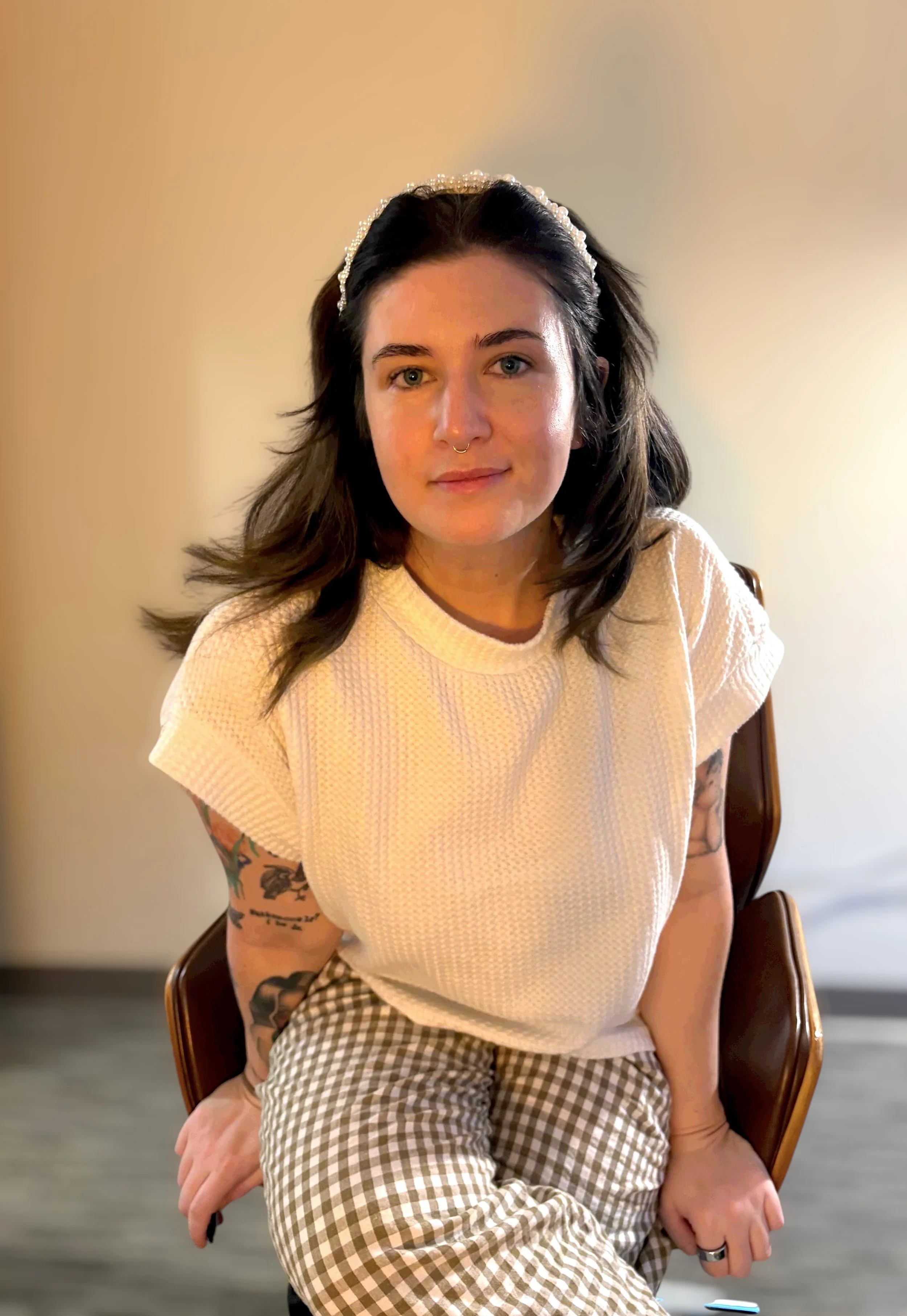 A young woman with dark hair, tattoos on her arms, wearing a white textured t-shirt and checkered pants, sitting on a wooden chair with a neutral background.