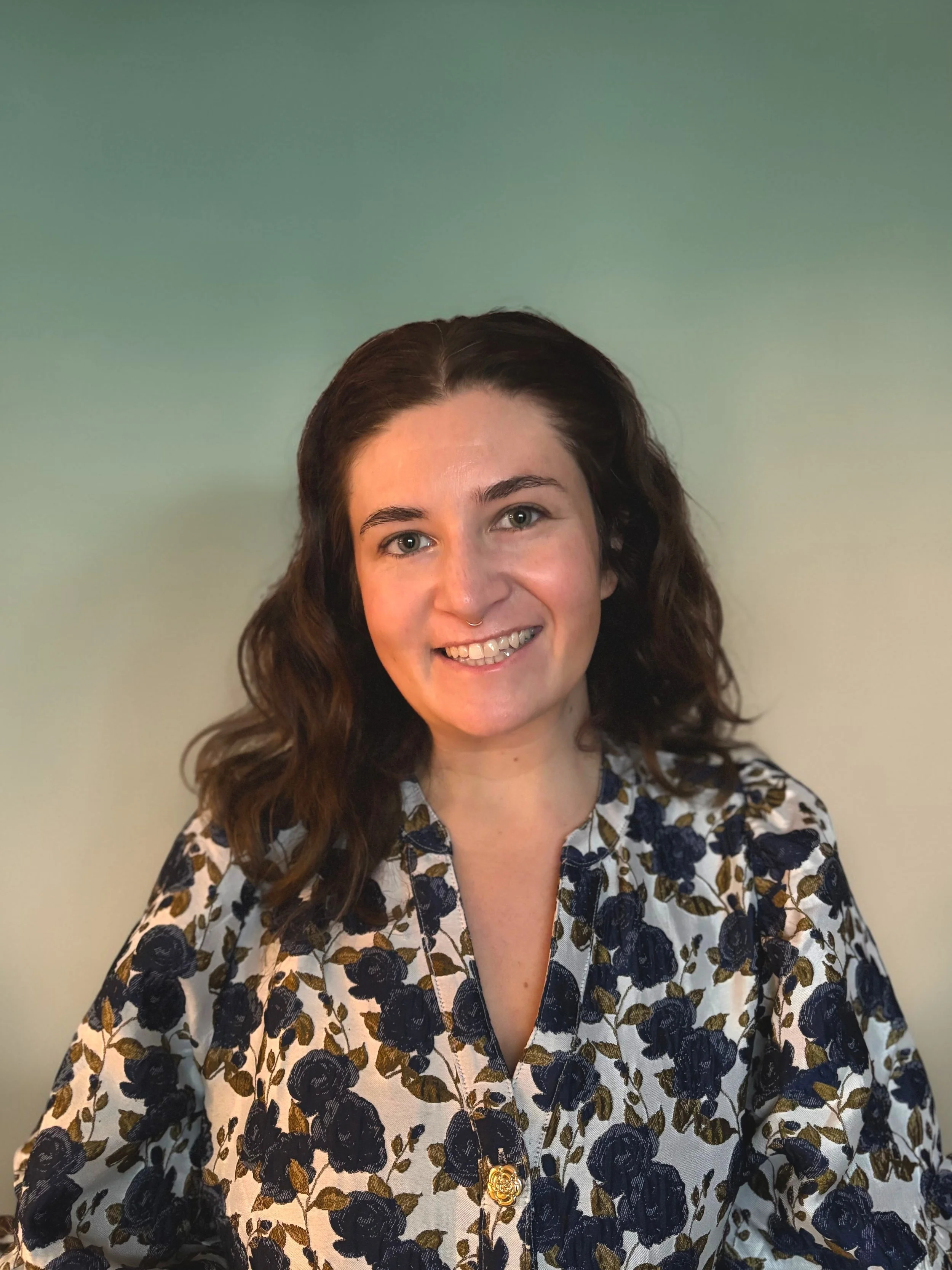 A woman with shoulder-length wavy brown hair, wearing a floral blouse with navy blue and white flowers, smiling at the camera against a gradient background.