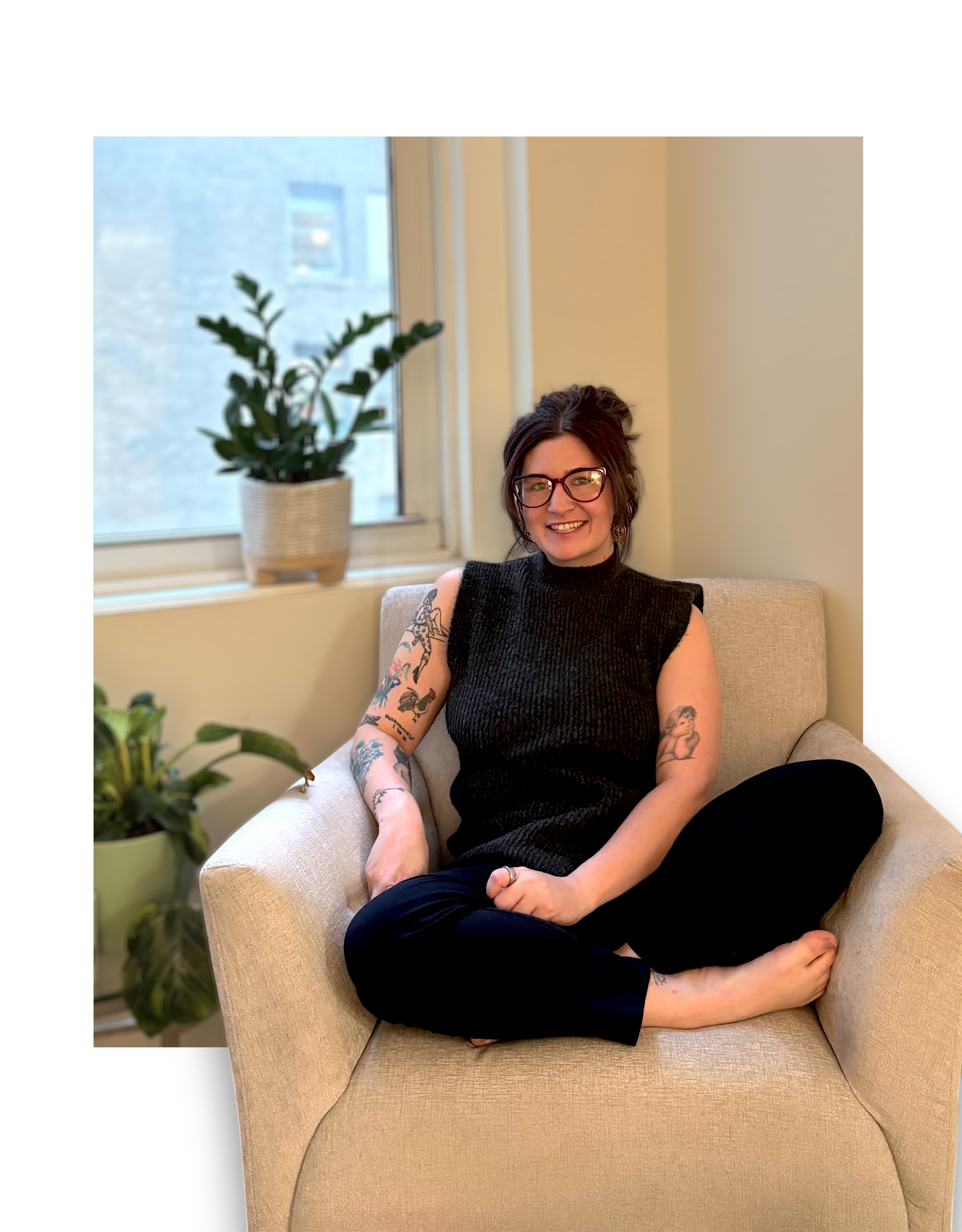 A woman with glasses and tattoos sitting on a beige armchair in an indoor space near a window with plants.