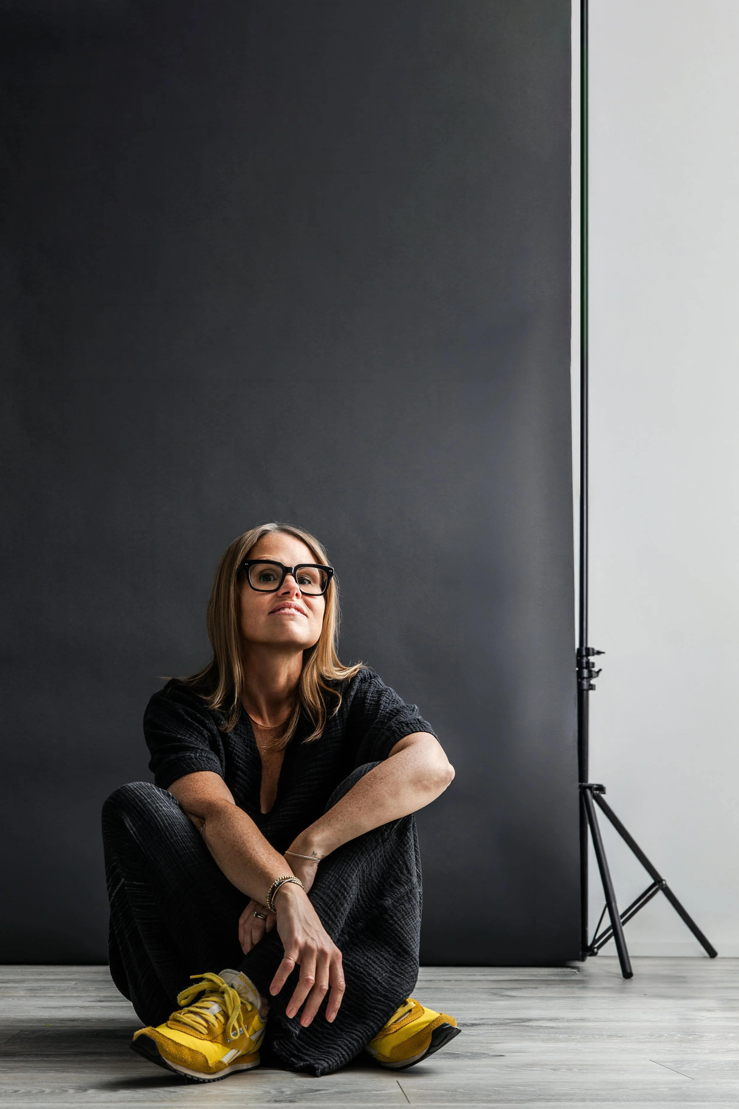 Black and white photo of Jen Ross sitting on the floor with her arms wrapped around her knees, wearing black pants, a black top, and yellow Reebok sneakers. The background includes a dark wall, a light wall, and a photography light stand.