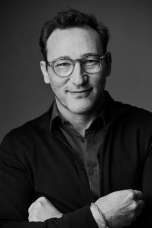 Black and white portrait of a Simon Sinek with glasses, short hair, wearing a dark shirt and smiling, against a plain background.
