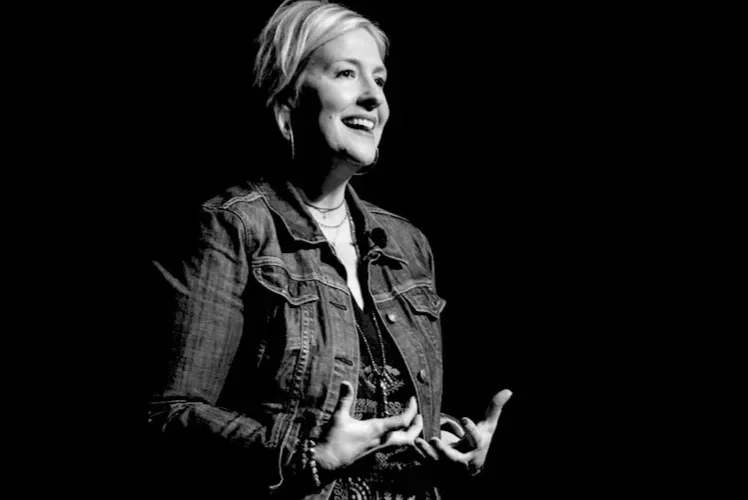 Black and white photo of a Brene Brown smiling, wearing a denim jacket, standing against a dark background.