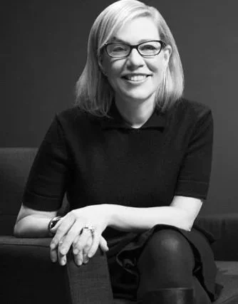 Black and white photo of a Debbie Millman with glasses, shoulder-length hair, smiling, wearing a dark shirt, sitting with arms crossed.