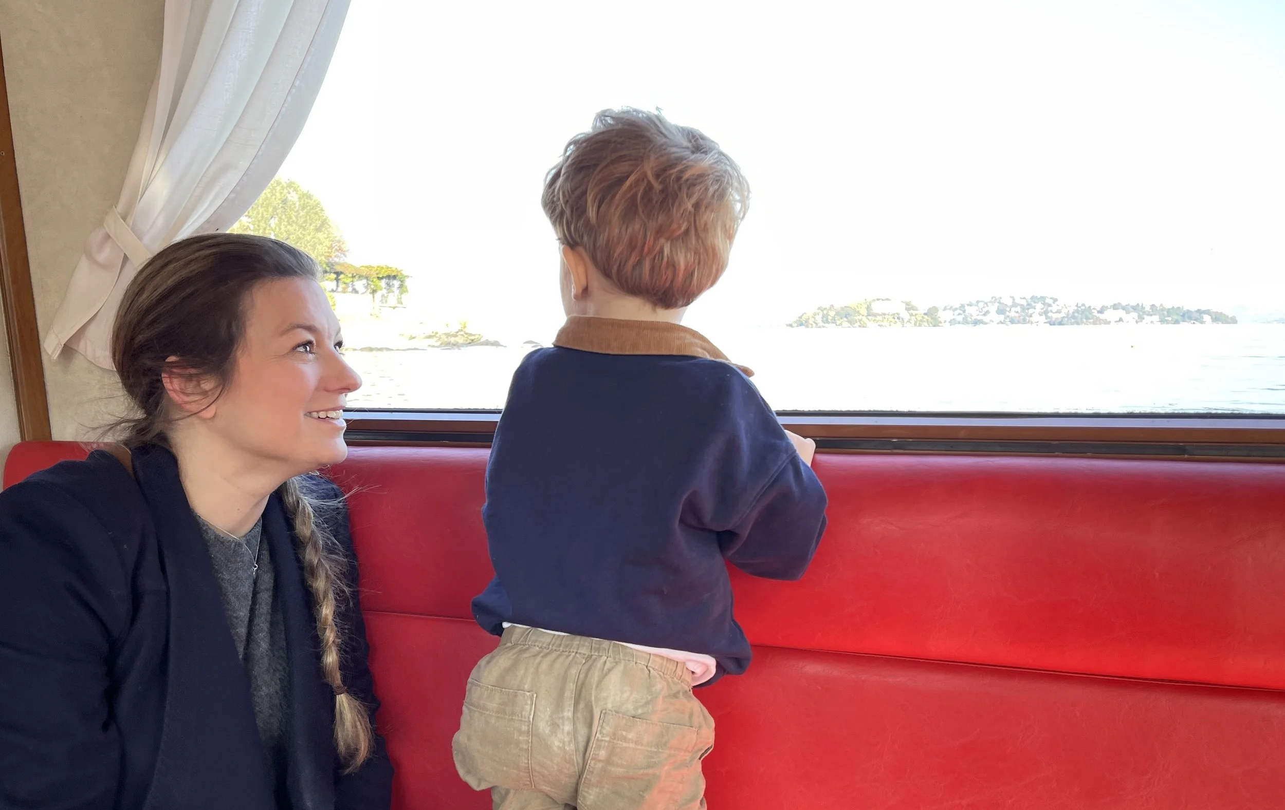 A woman with long brown hair, smiling and sitting on a red cushioned seat, looking at a young boy who is standing and looking out of a window onto a body of water and distant land.