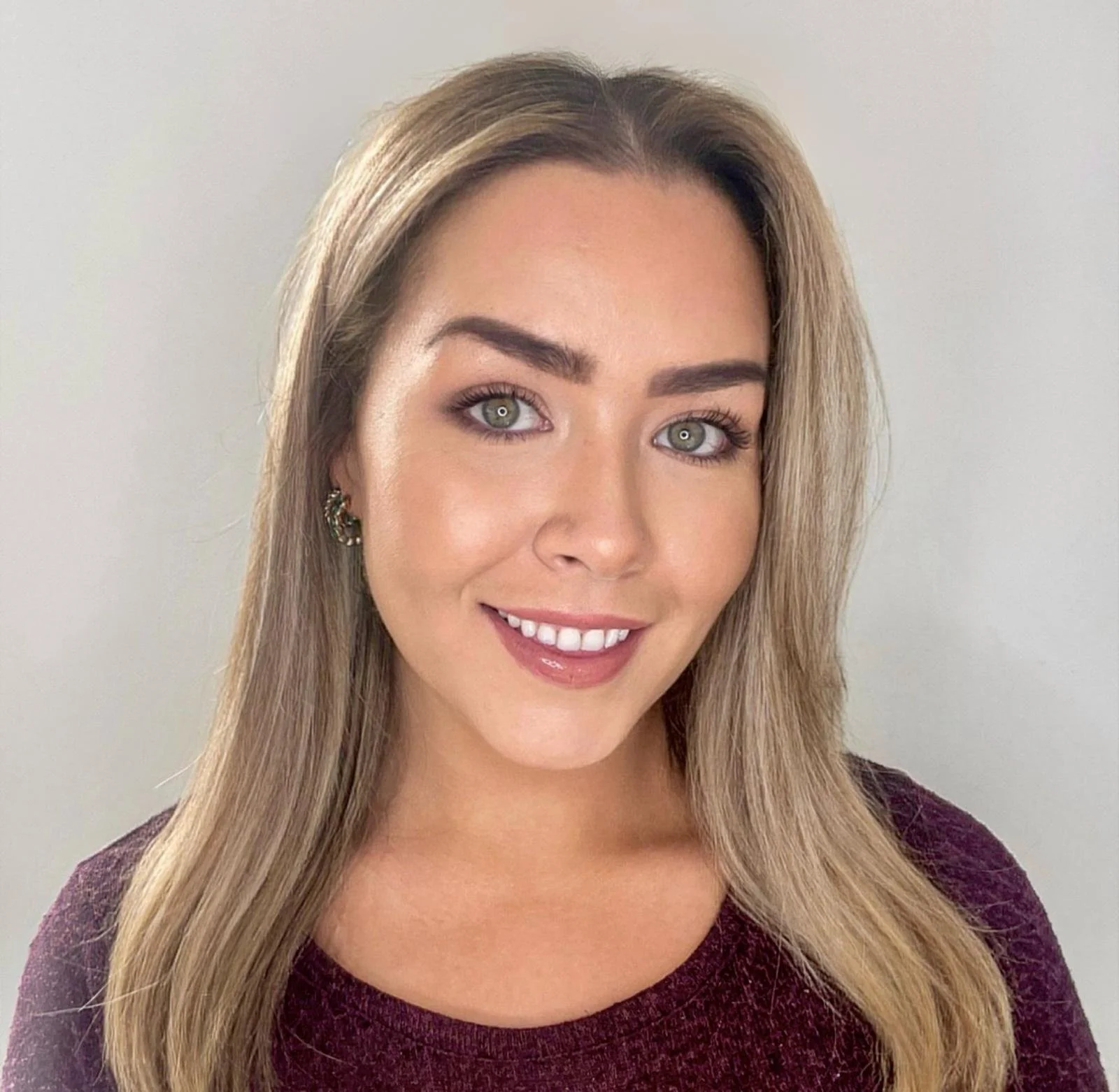 A woman with long blonde hair, wearing a maroon top and earrings, smiling against a plain light gray background.