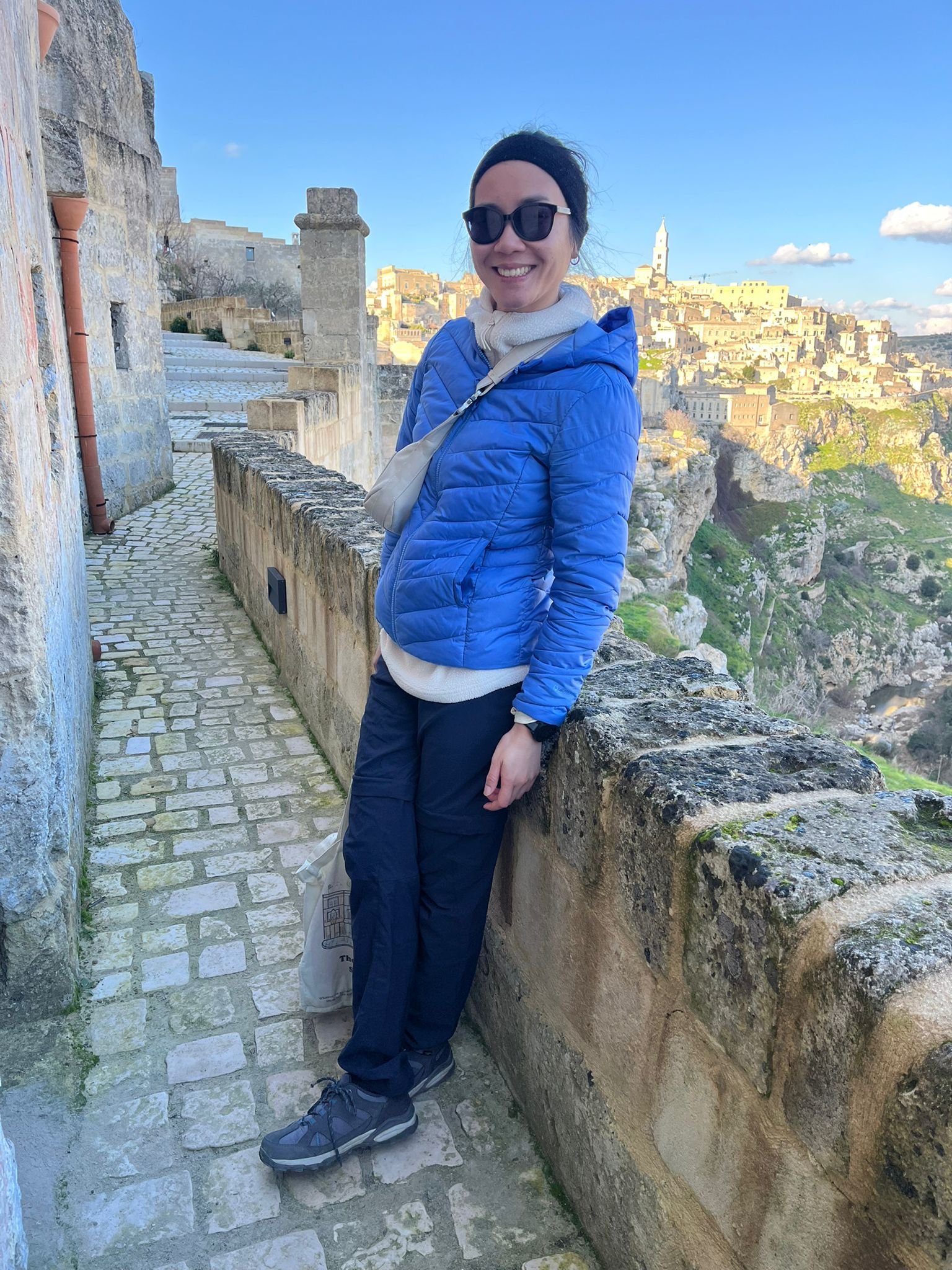 Woman in sunglasses and blue jacket standing on a cobblestone pathway with a stone wall, with a scenic historic cityscape in the background on a clear day.