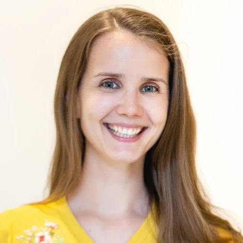A young woman with long red hair, blue eyes, and fair skin, smiling at the camera, wearing a yellow top with floral embroidery, against a plain light-colored background.