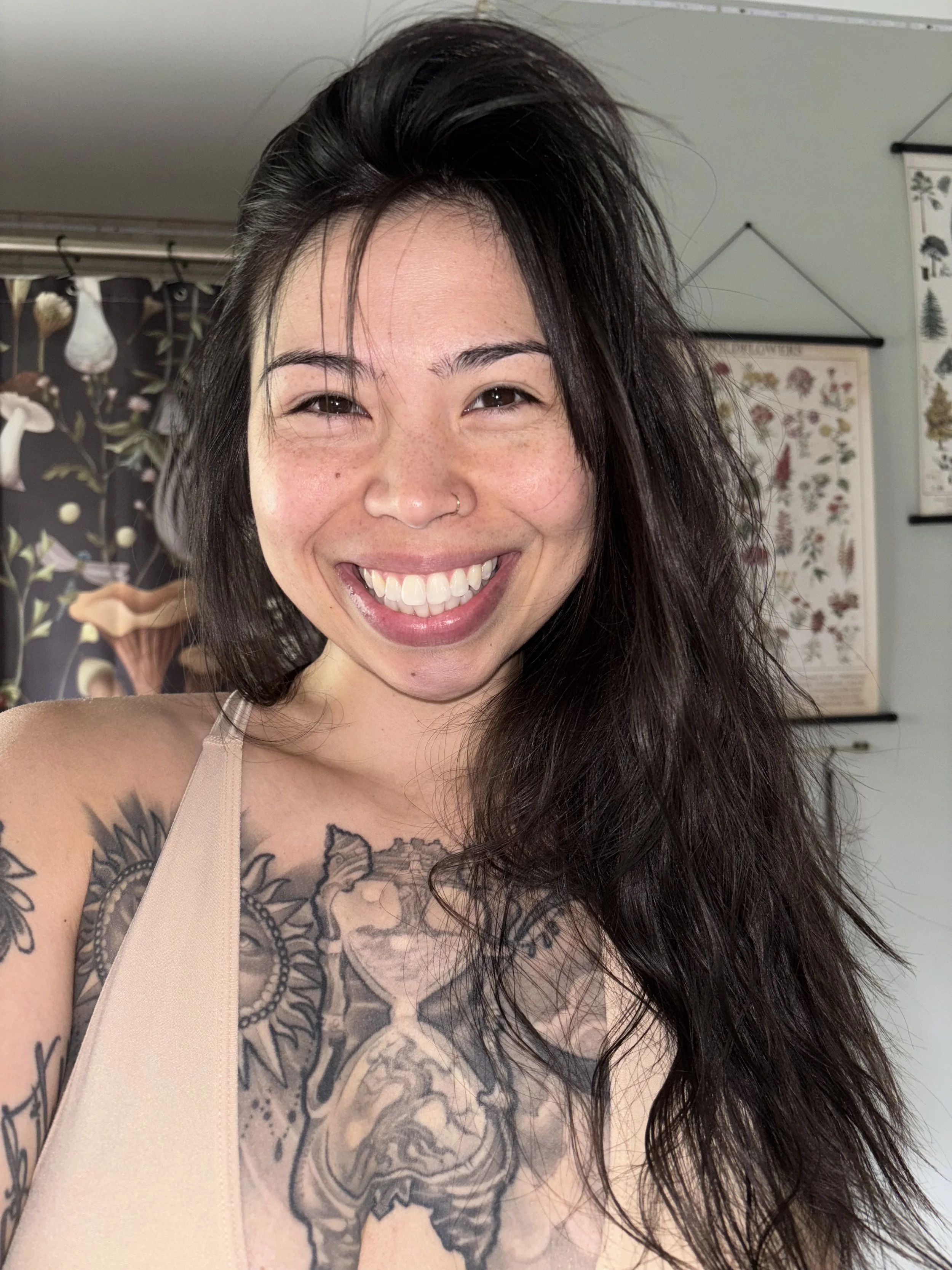 Close-up of a smiling woman with dark, wavy hair, a septum piercing, and tattoos on her chest and shoulder, in an indoor setting with wall posters and plants in the background.