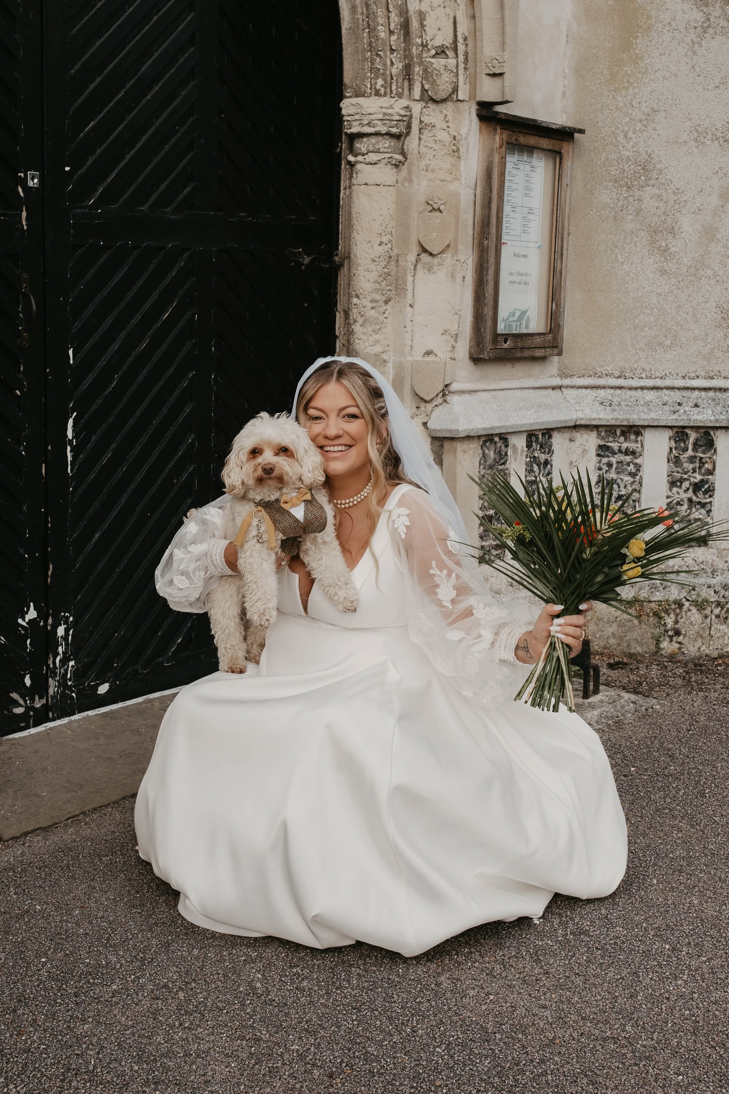 A woman in a wedding dress sitting on the ground outside, holding a bouquet of flowers in one hand and a small dog in her lap. She is smiling and wearing a bridal veil. The dog is dressed in a small outfit, and they are in front of a historic stone building with a black door and an outdoor sign.