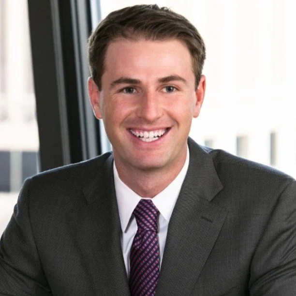 A smiling man in a dark suit, white shirt, and purple tie, sitting in front of a window in an office environment.