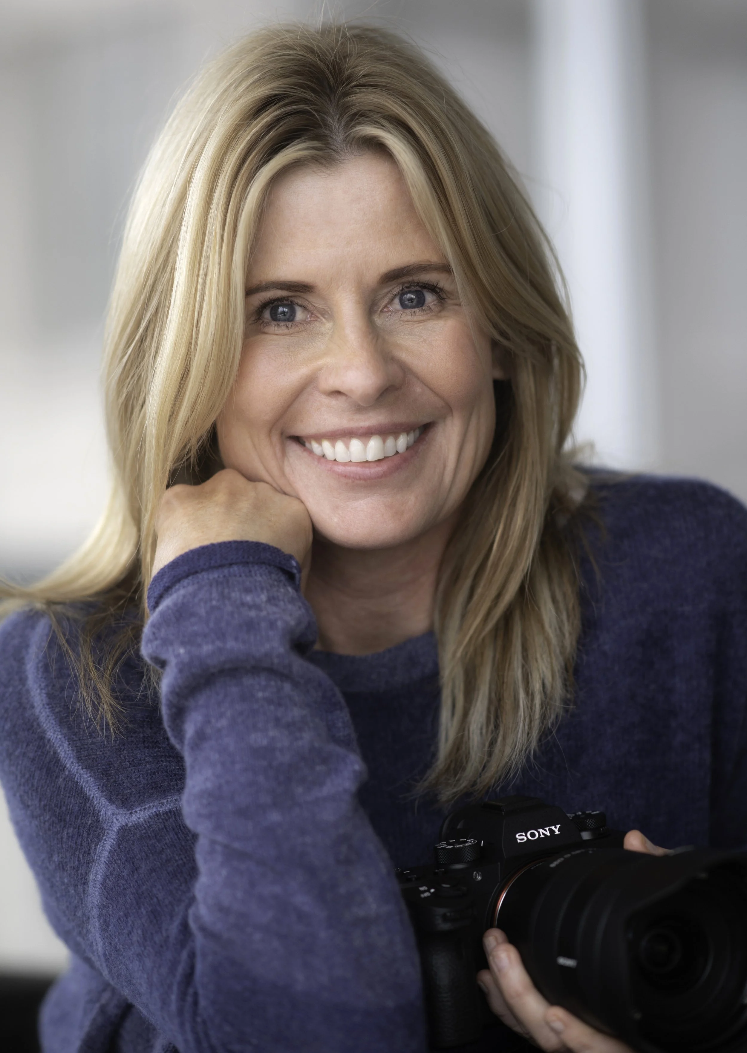 A woman with blonde hair wearing a blue sweater, smiling and holding a Sony camera.