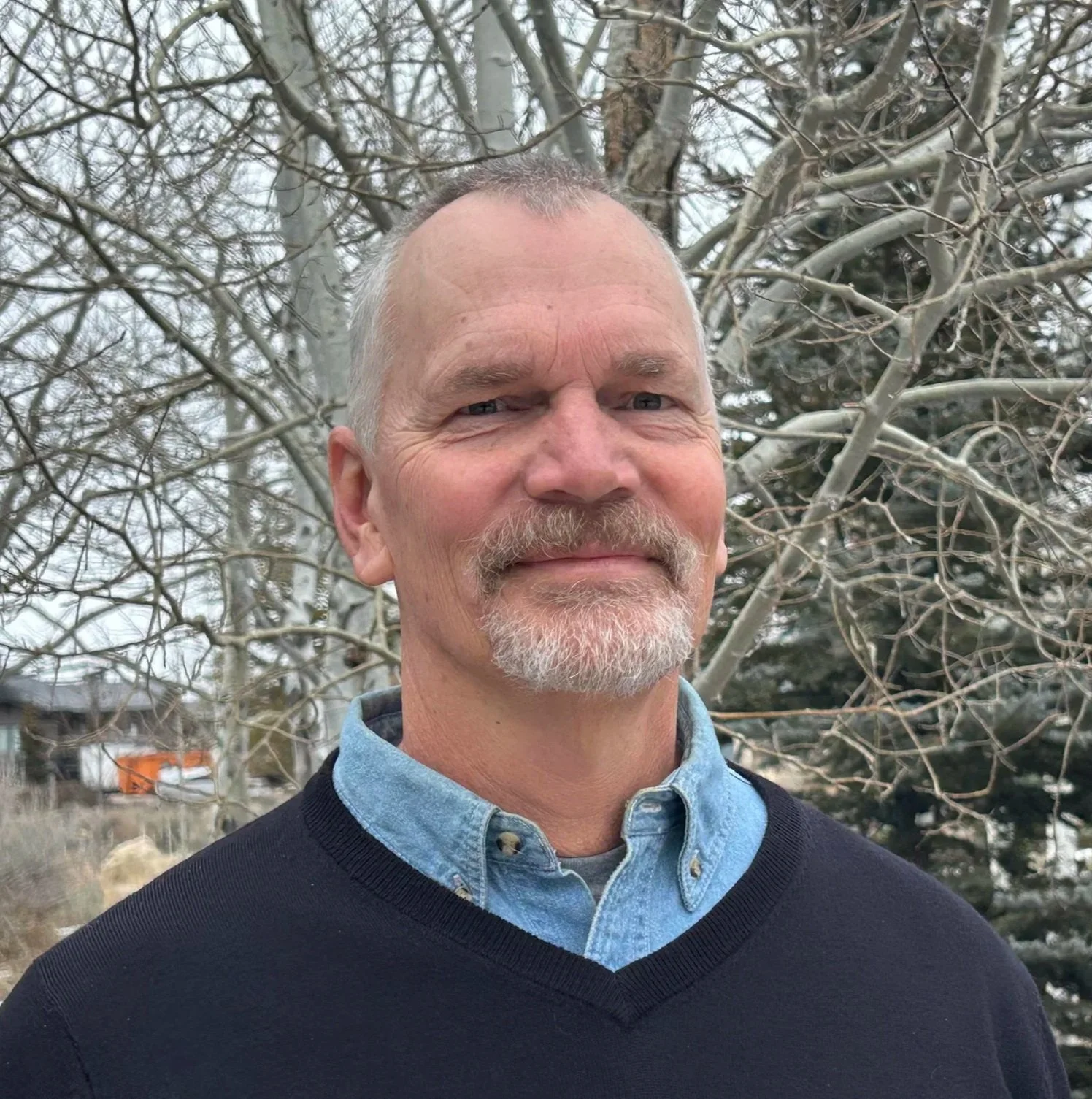A middle-aged man with short gray hair, a goatee, and blue eyes standing outdoors near leafless trees, wearing a blue denim shirt under a black sweater.