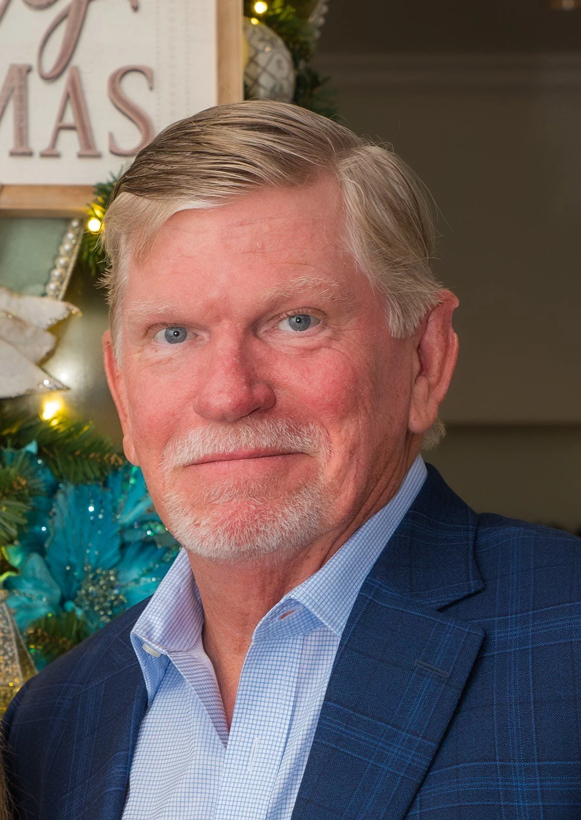 Portrait of a mature man with gray hair and a beard, wearing a blue checkered suit and light blue shirt, standing in front of a decorated Christmas tree.