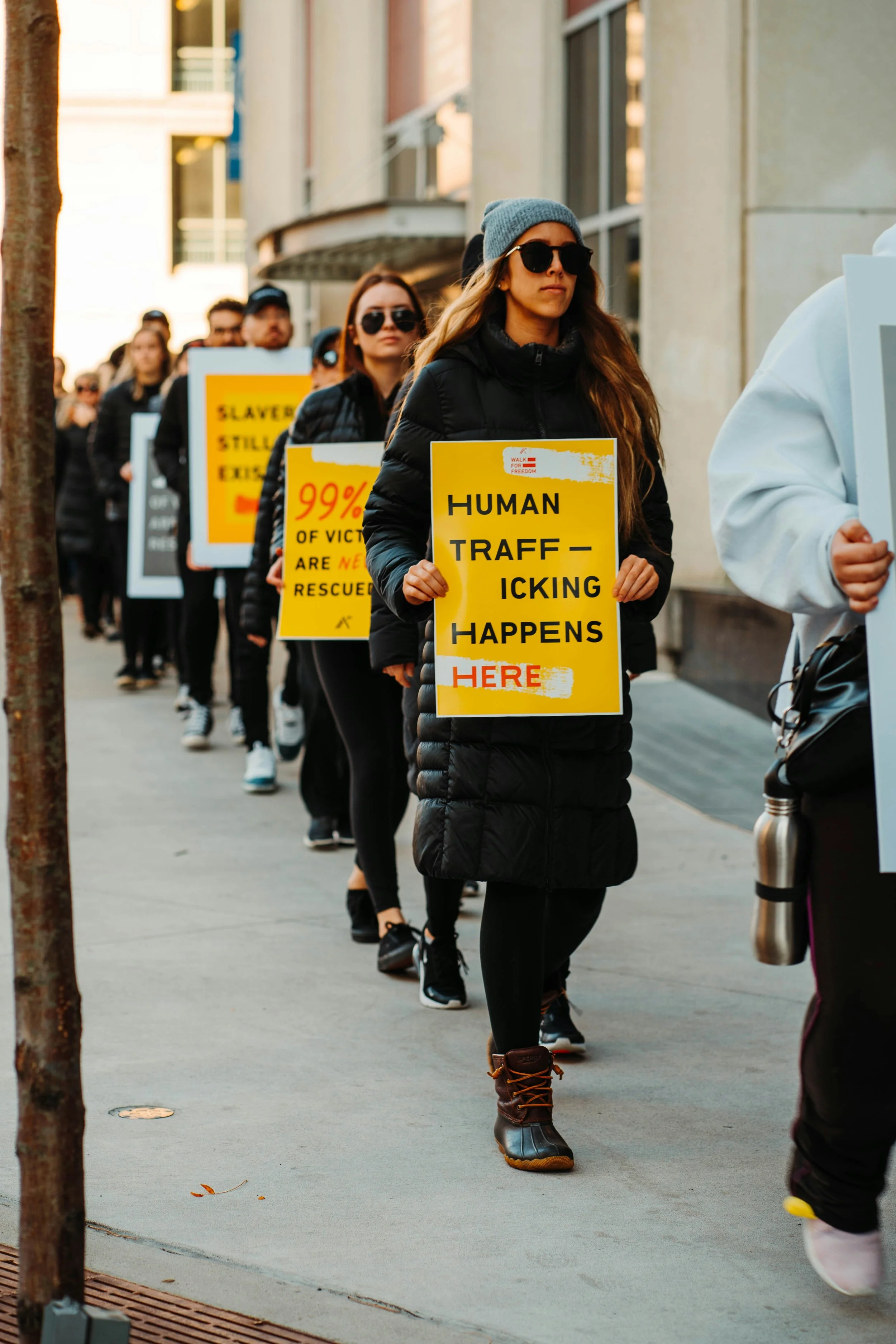 Line of people holding protest signs about human trafficking, with the woman in front wearing a black coat, gray beanie, sunglasses, and brown boots.