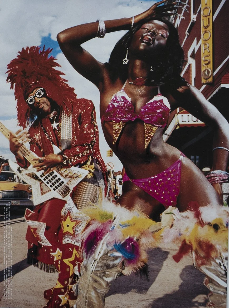 A woman and a man performing a parade in colorful costumes on a street with buildings and cars in the background. The woman is wearing a revealing pink and gold outfit with feathered accessories, and the man is dressed in a sequined red costume with a large feathered headdress, playing a guitar.