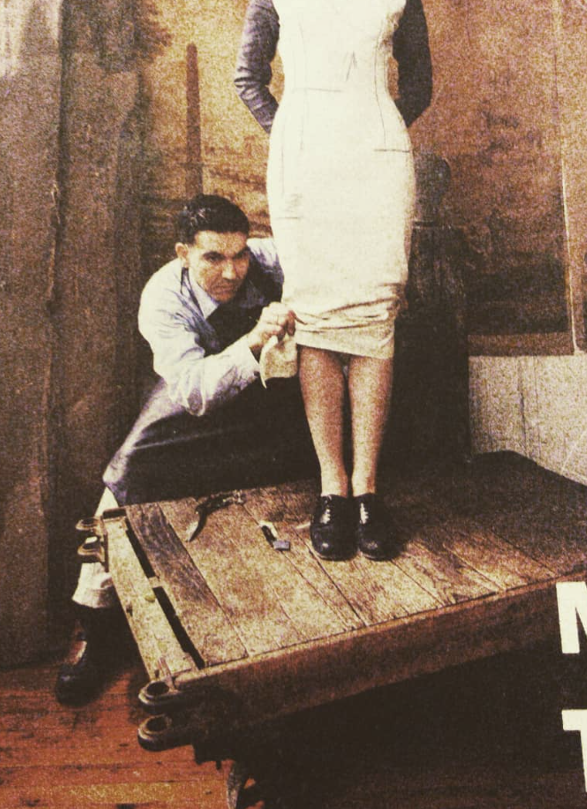 A man crouching beside a woman standing on a wooden platform, with the woman wearing a beige dress and black shoes, partially lifting the dress revealing her legs. The man is holding the hem of her dress, and the setting appears to be indoors with wooden and textured walls.