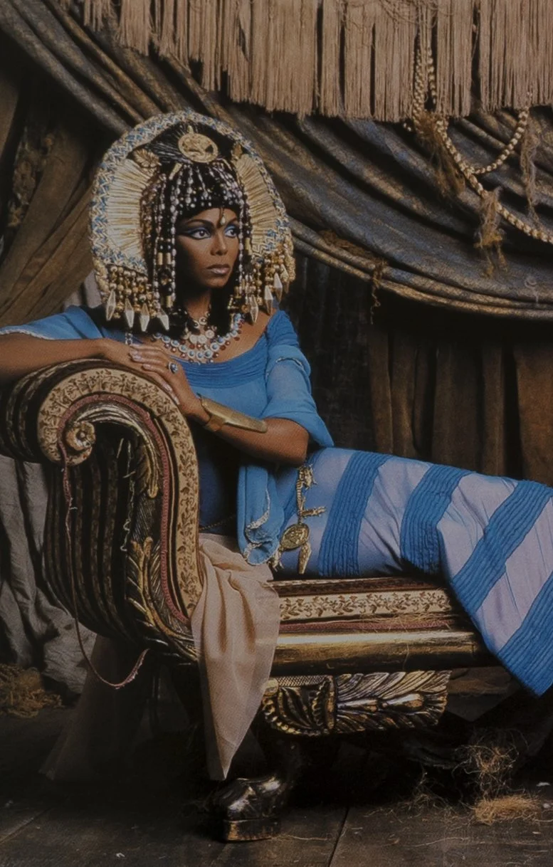 A woman dressed in ancient Egyptian attire, wearing a large ornate headdress, blue dress, and jewelry, reclining on an ornate chaise lounge with a wooden backdrop.