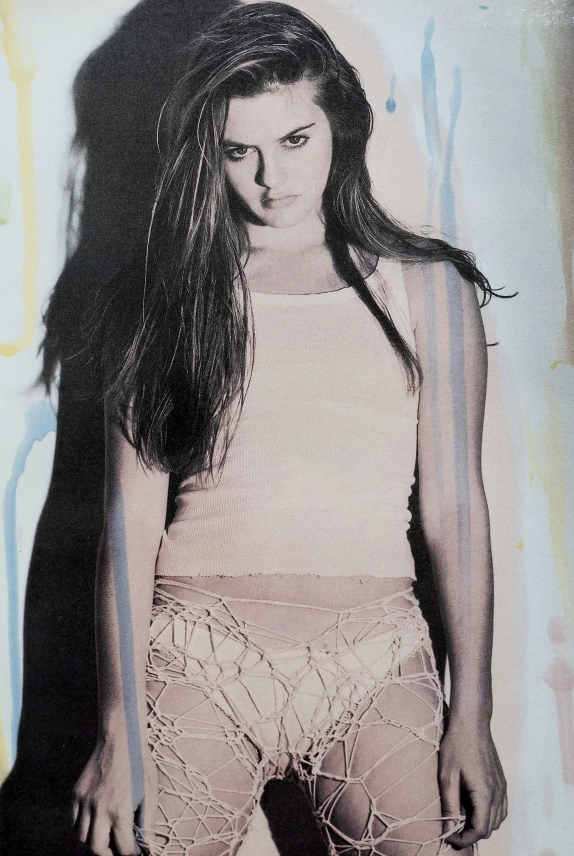 Black and white photo of a young woman with long, wavy hair, wearing a sleeveless top and fishnet tights, standing against a wall with her shadow cast to her left.