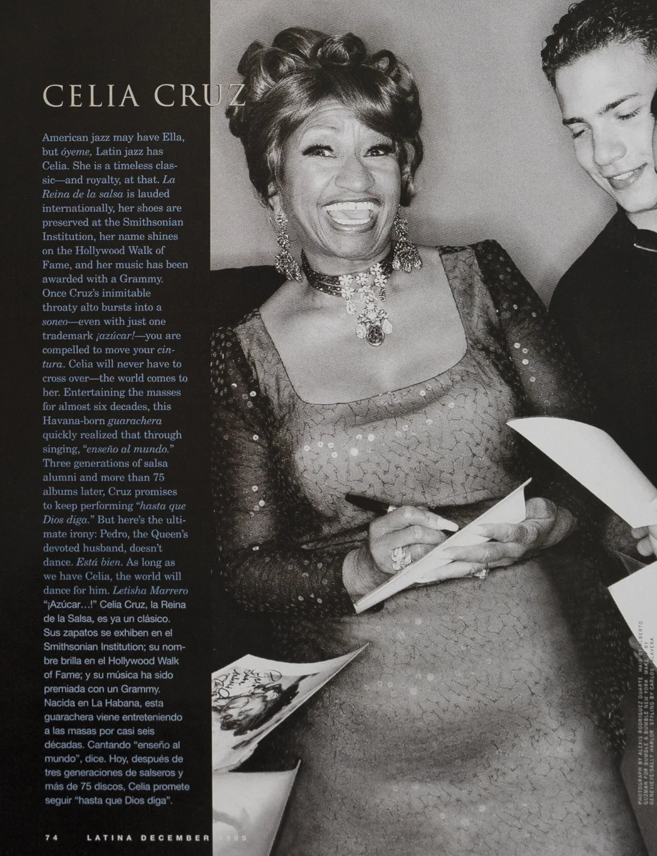Black-and-white photo of a woman with short curly hair smiling, dressed in a sparkly dress, wearing jewelry, and holding a pen while signing autographs. Part of another person is visible to her right.