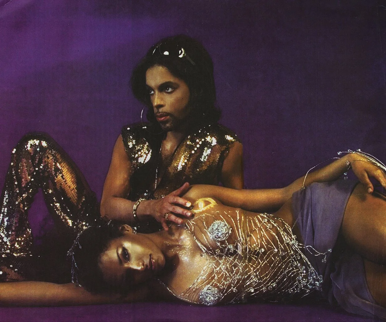 Two individuals dressed in shiny, glittery clothing are lying on purple surface, with one person resting their head on the other's chest. The scene has a glamorous, artistic vibe.