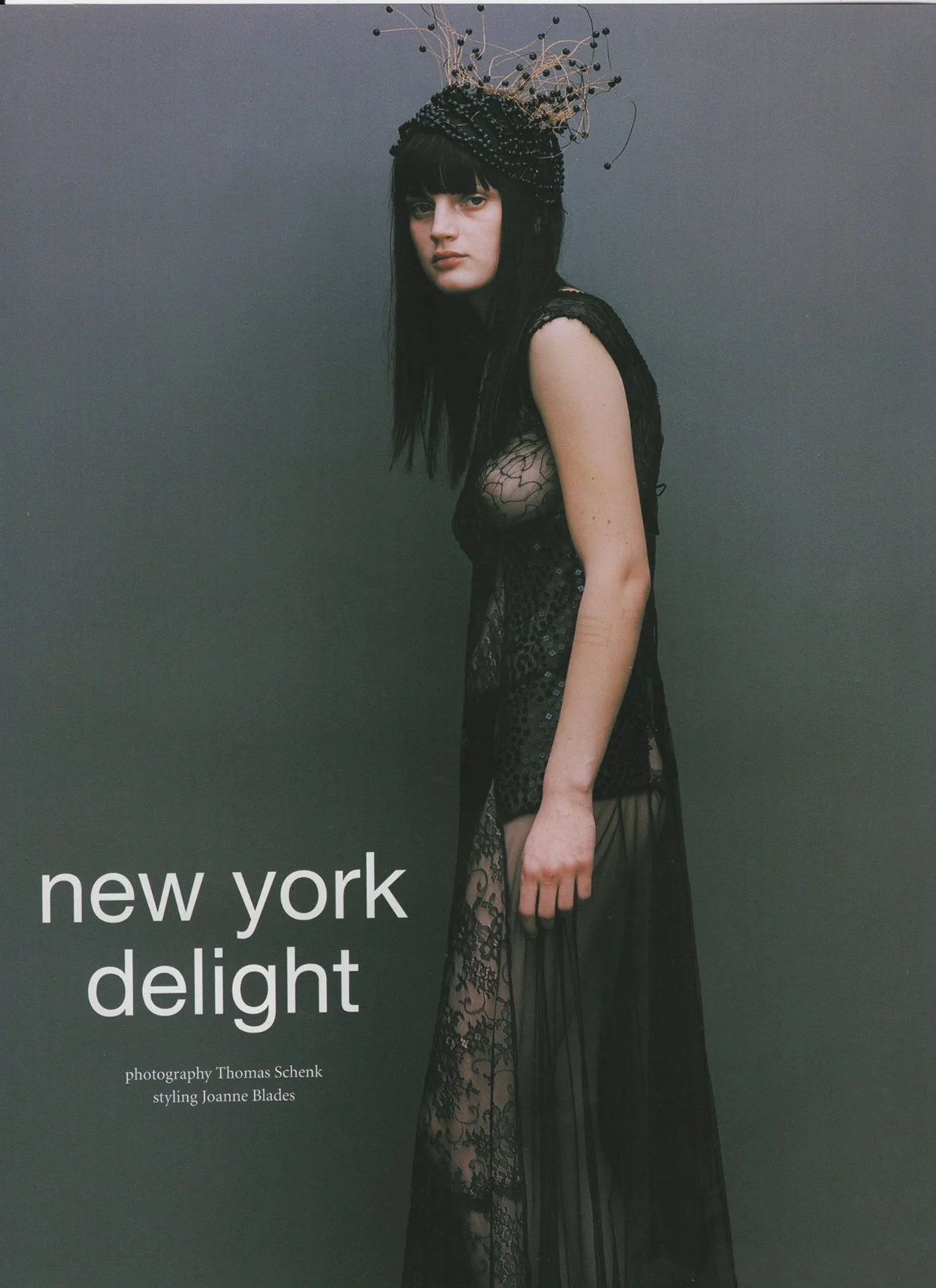 A woman with black hair and a dark headpiece with wire and beads wears a sheer black lace dress and stands against a gray background with text 'new york delight' and photography credit.