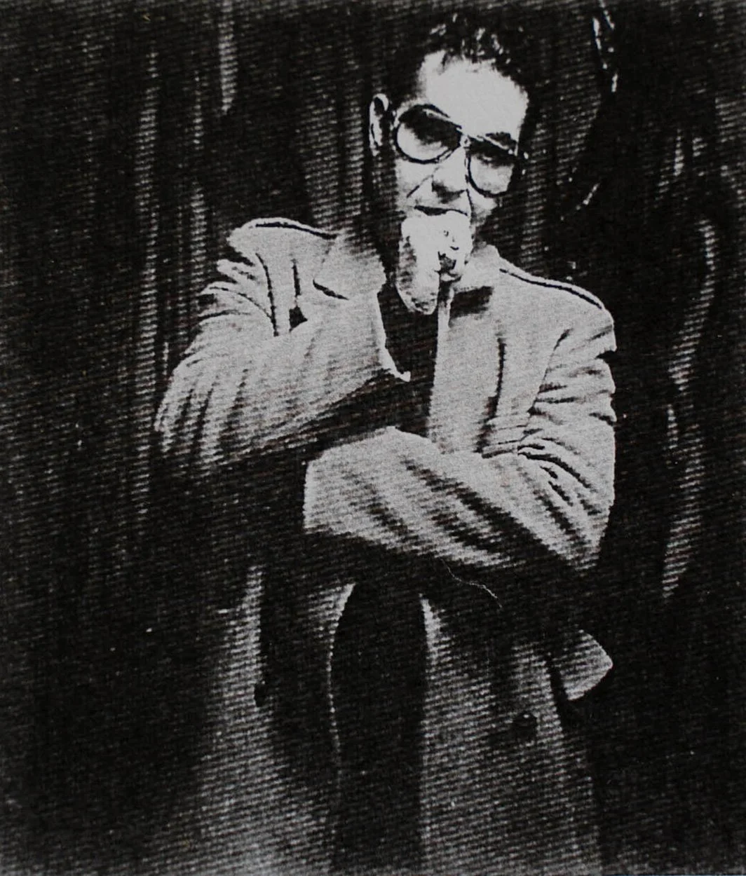 Black and white photo of a person with glasses and short hair, wearing a jacket, standing in front of a curtain, with a hand on their chin, making a thoughtful or contemplative gesture.