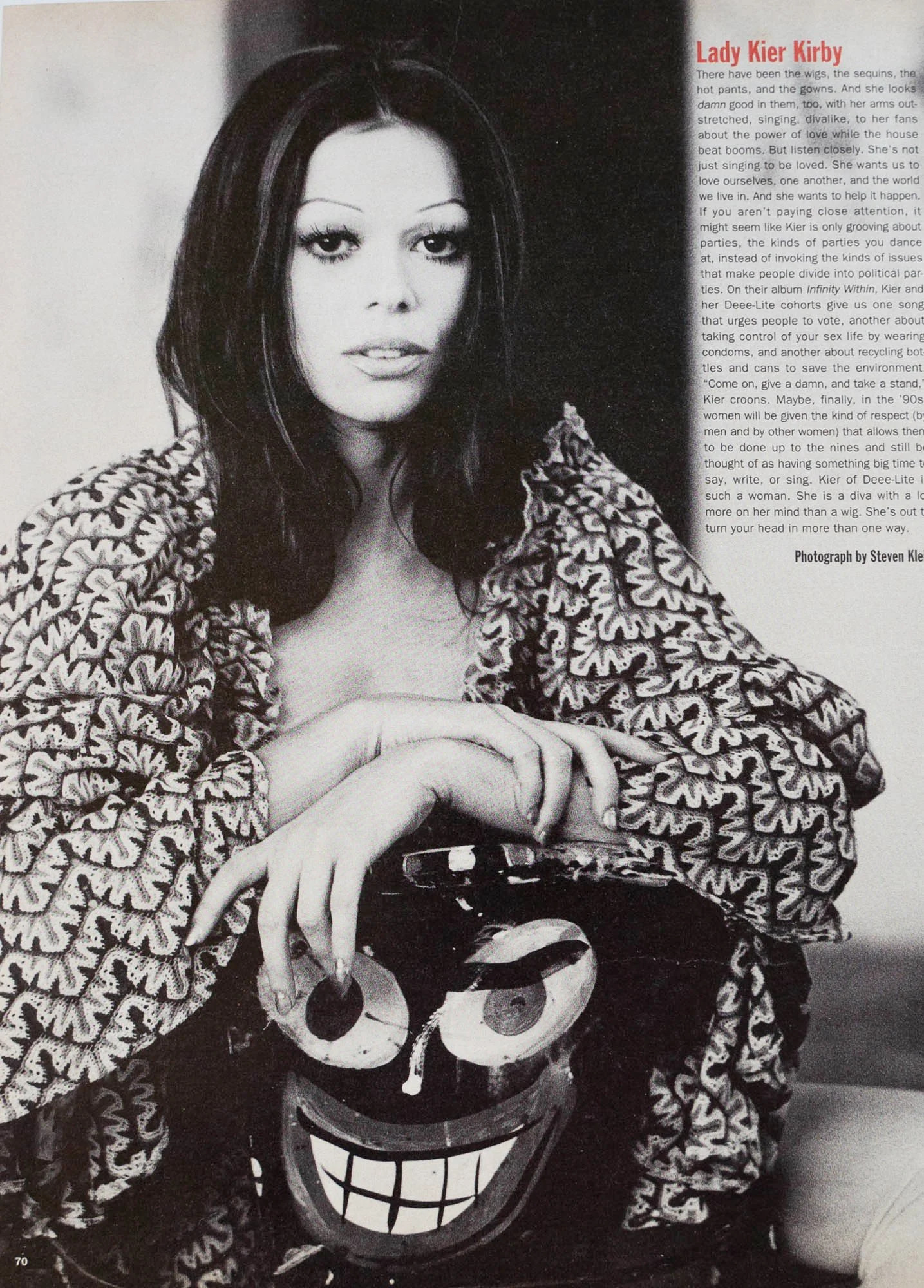 Black and white photograph of singer Lady Kier Kirby posing with a painted smiling face mask, wearing a patterned jacket, with a column of text on the right side of the image.