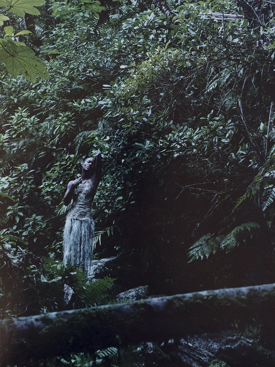 A woman in a garden surrounded by dense green foliage, standing on a small waterfall or stream, with her hand behind her head, wearing a textured dress.