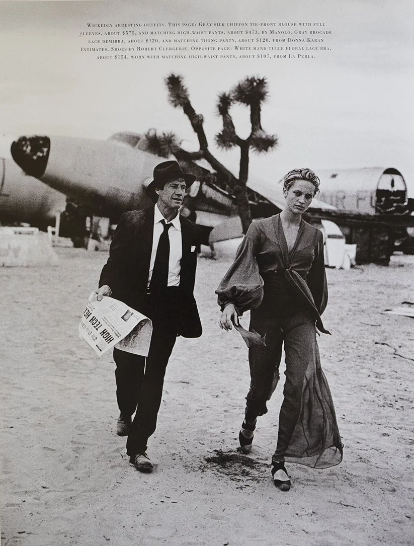Black and white photo of a man and a woman walking through a desert, with a helicopter and a josh tree in the background. The man is wearing a suit and hat, holding a newspaper, while the woman is dressed in a long, flowing dress and high heels.