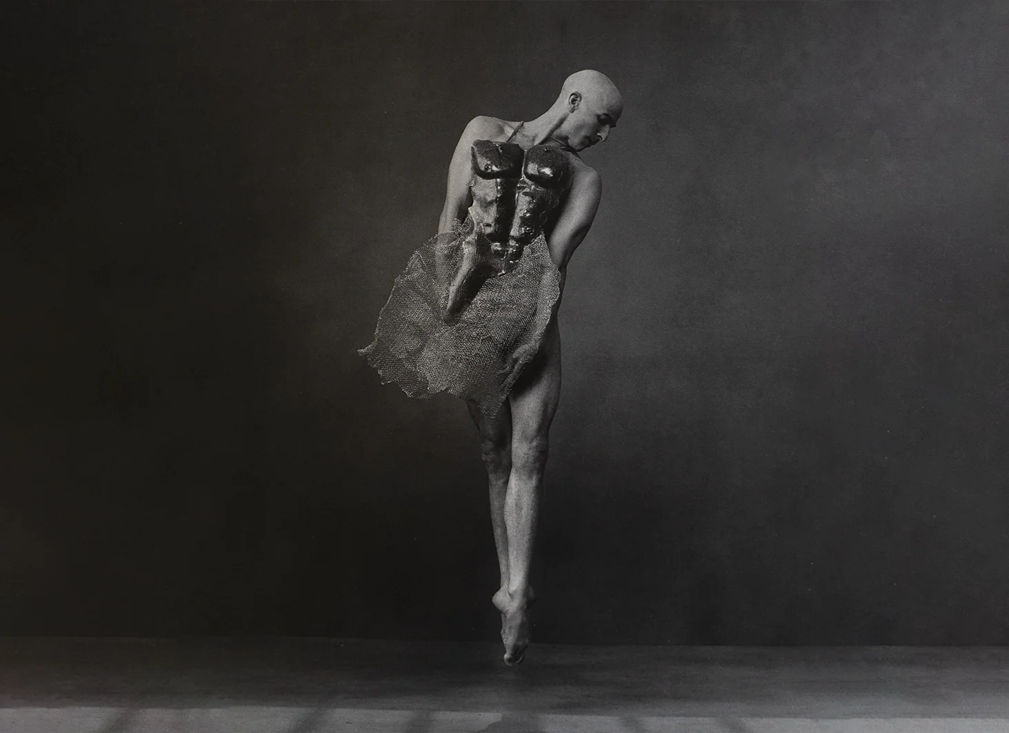 A black and white artistic photo of a human figure with a bald head, wearing a textured skirt, and holding a dark mask in front of their chest, standing on tiptoe against a dark background.