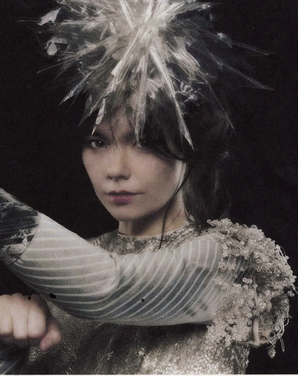 A woman wearing a metallic, textured dress with ornate embellishments on the shoulders, and a large, spiky, transparent headpiece with silver accents, striking a confident pose against a dark background.