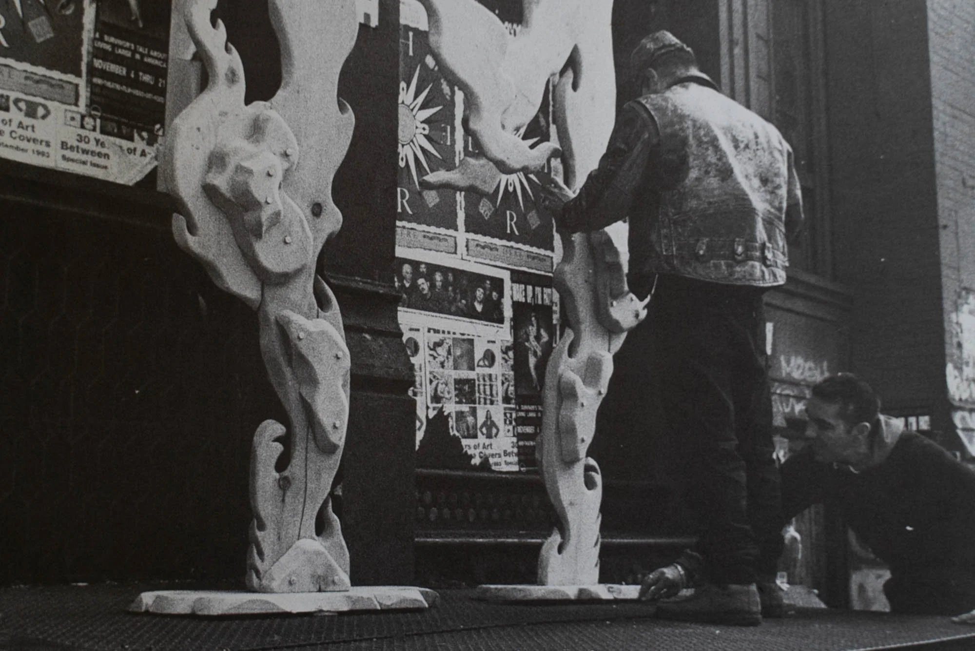 Two men working on wooden sculpture pieces, one standing and another kneeling, in an indoor workshop. The sculptures have ornate, curving designs and are placed on a work table.