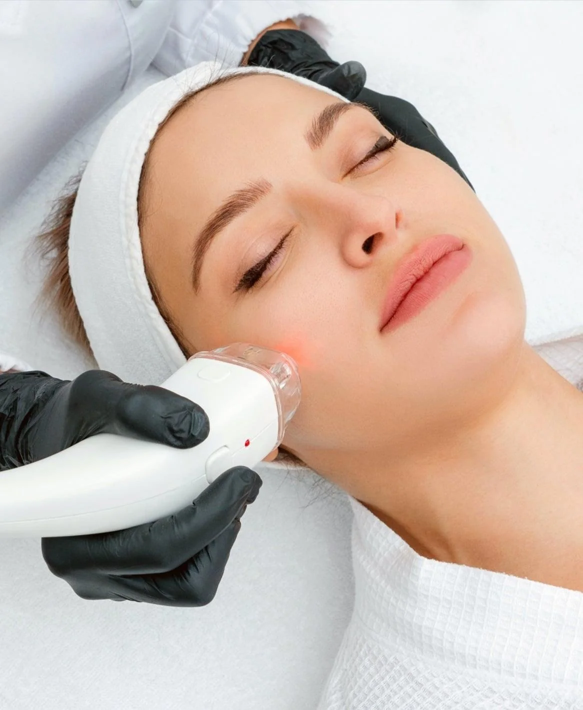 A woman receiving a facial treatment with a specialized device applied to her cheek in a spa or clinic setting.