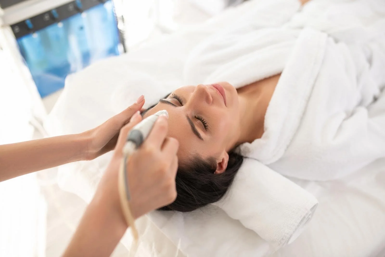 Woman receiving a facial treatment in a clinic setting, laying down with eyes closed, with a towel wrapped around her head.
