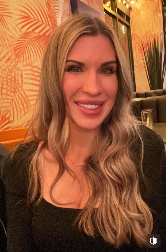 A woman with long wavy blonde hair smiling at a restaurant or bar with warm lighting and tropical patterned wallpaper in the background.