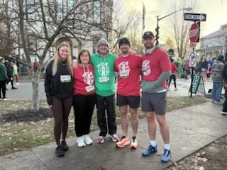 Benjamin Ligan stands with four others at an outdoor race event, all wearing matching red race shirts, gathered on a sidewalk near street signs.