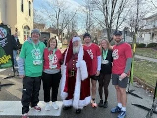 Benjamin Ligan and four others pose with a person dressed as Santa Claus at an outdoor race event, all smiling and wearing red race shirts with medals.