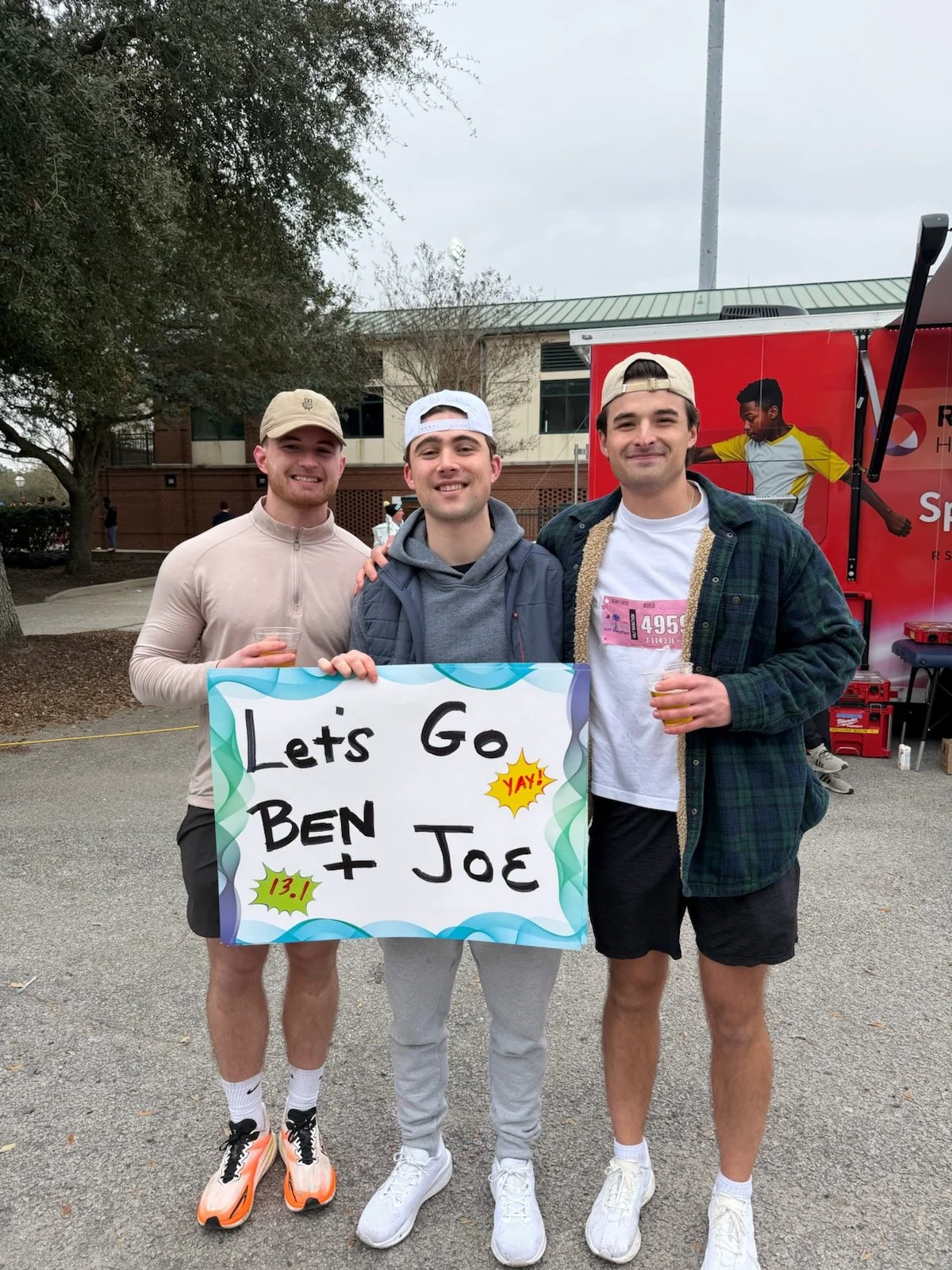 Benjamin Ligan stands with two friends at a half marathon event, smiling and holding a handmade sign that reads 'Let's Go Ben + Joe 13.1,' with a festive outdoor backdrop.