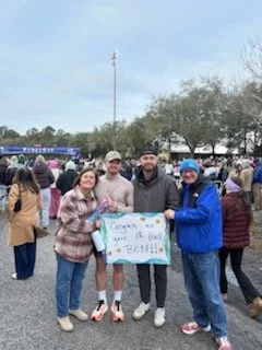 Benjamin Ligan celebrates after a half marathon with a group of four friends and family members, one of whom is holding a sign reading 'Congrats on your 1st Half BEN!!'
