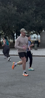 Benjamin Ligan running during a race, wearing a beige long-sleeve shirt, black shorts, and bright orange sneakers, with trees and other runners visible in the background.