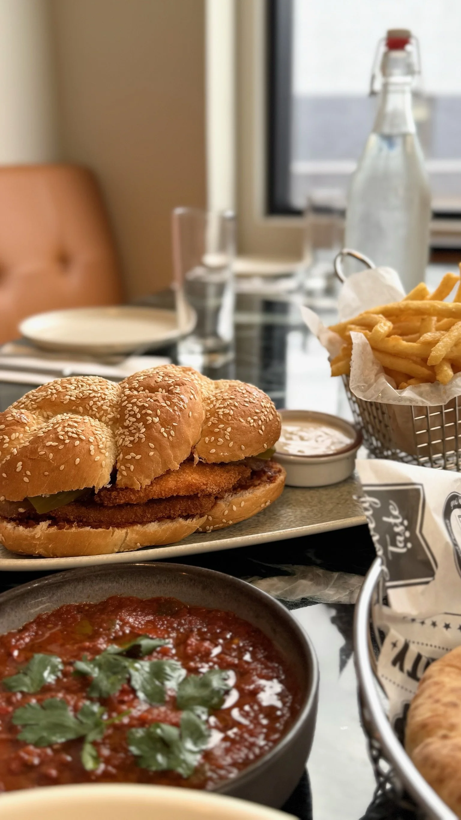 Close-up of a basket of French fries, a sandwich with fried chicken, lettuce and pickles, a bowl of tomato-based sauce garnished with cilantro, and a small container of dipping sauce on a restaurant table.