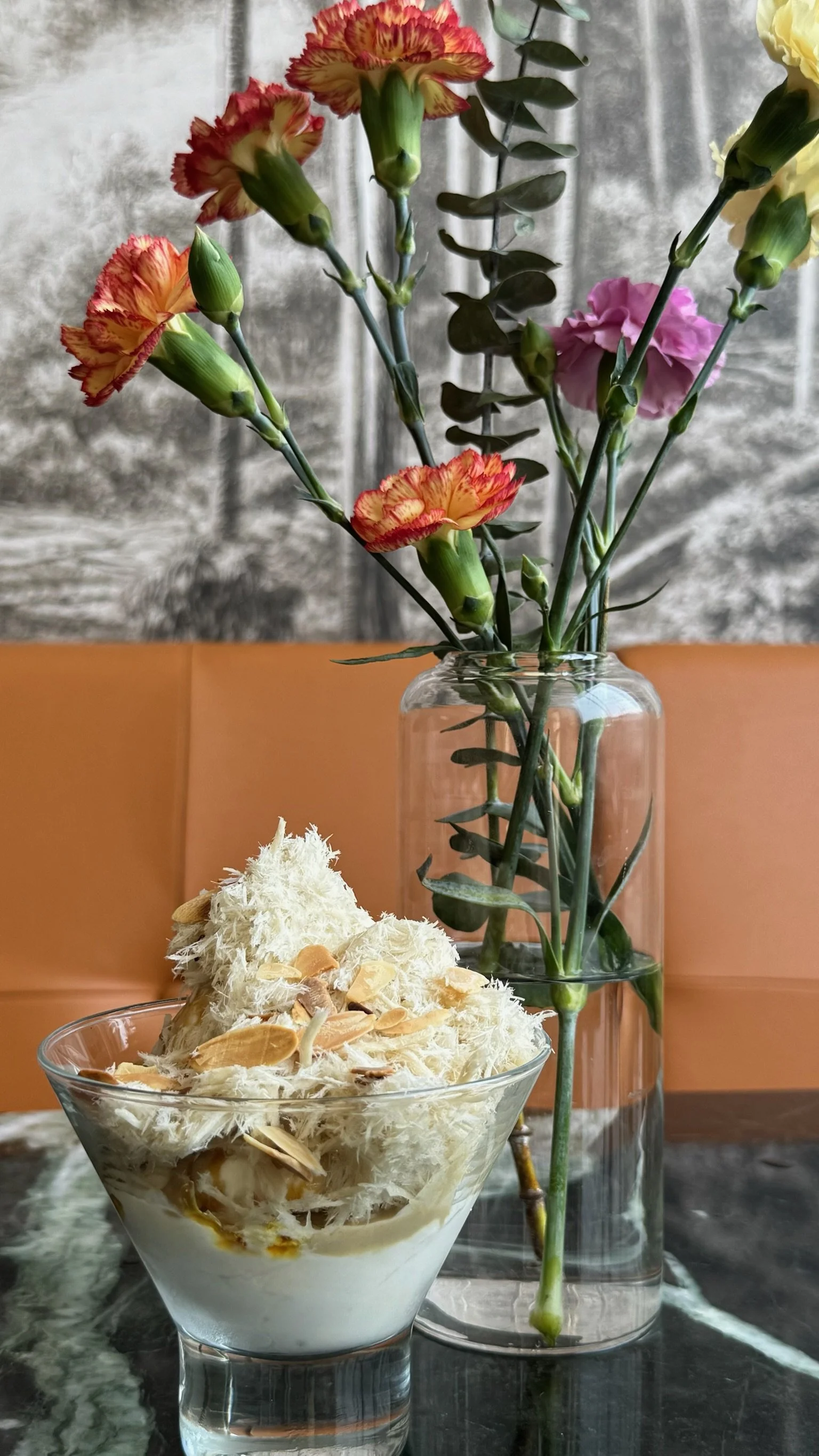 A glass bowl filled with shredded white and beige food, topped with almond slices. Behind the bowl, there is a tall glass vase with carnations in red, yellow, and pink, placed on a dark marble table.