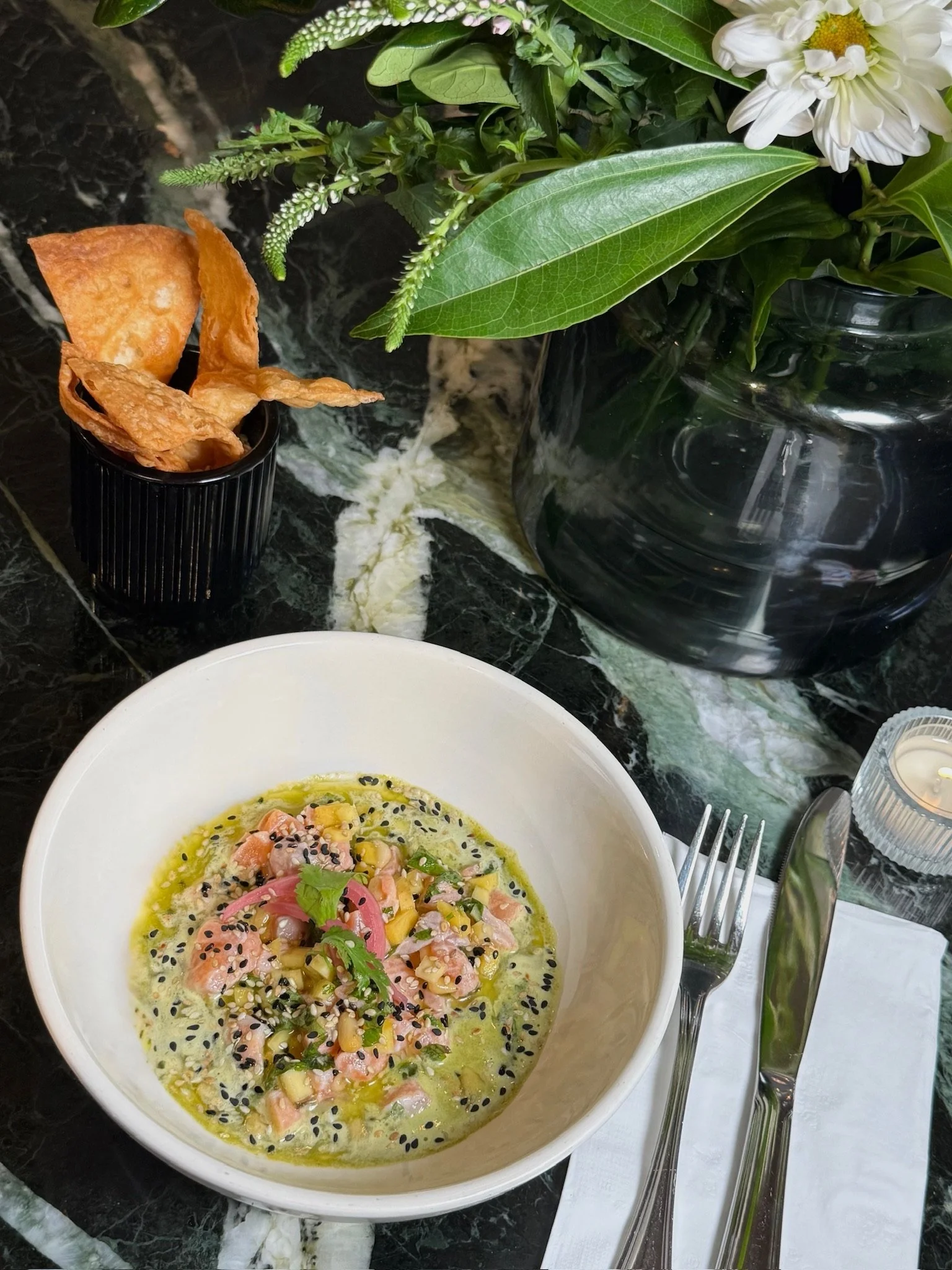 A dining table with a white bowl of poke including chopped fish, vegetables, and black sesame seeds. A fork and knife are placed on a white napkin beside the bowl. To the top right, there is a large black vase with green leaves and white flowers. To 