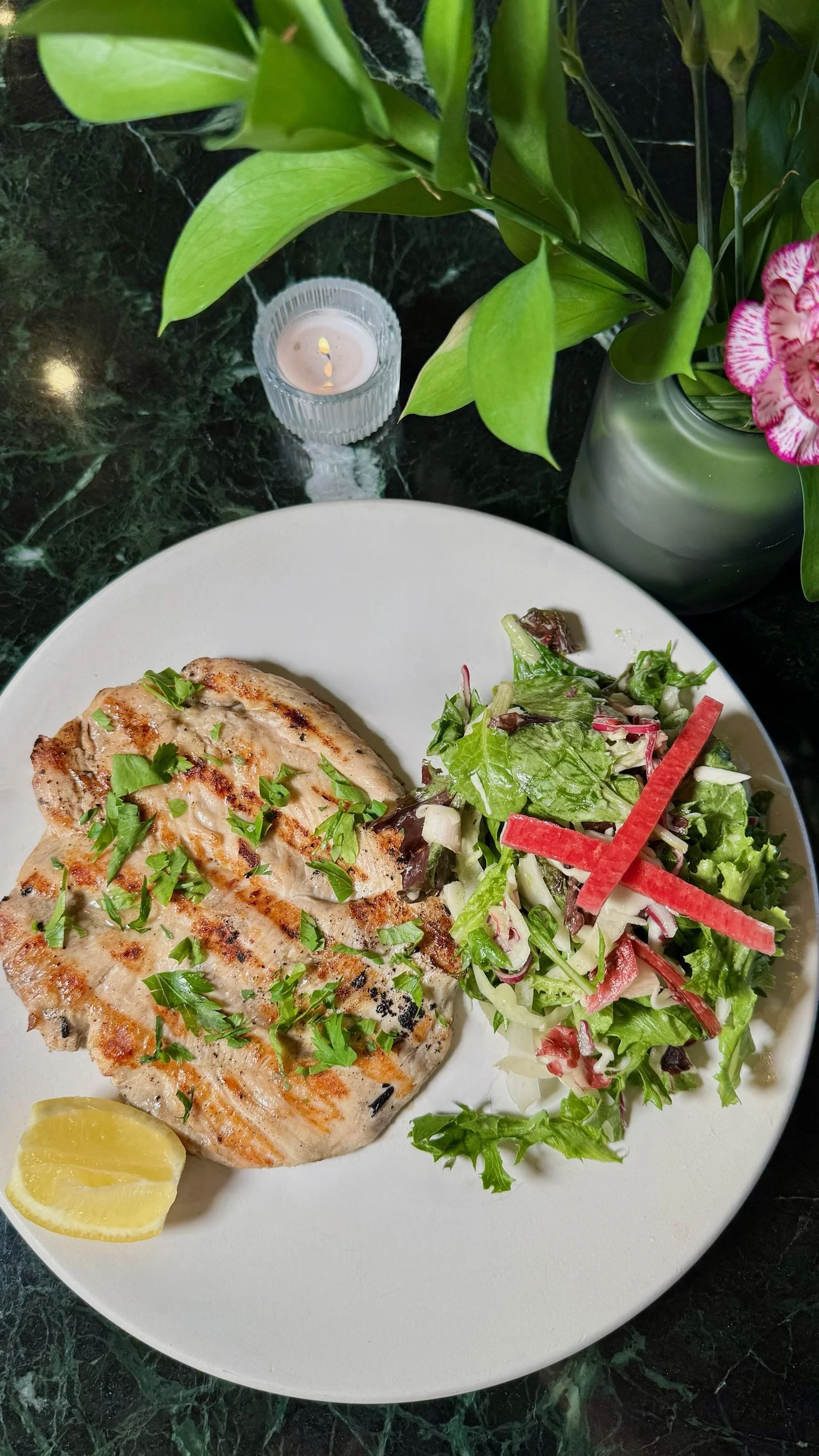 A plate with grilled chicken topped with herbs, a lemon wedge, and a side salad with mixed greens and slices of red bell pepper. A small candle, green leaves, and pink flowers are in the background.