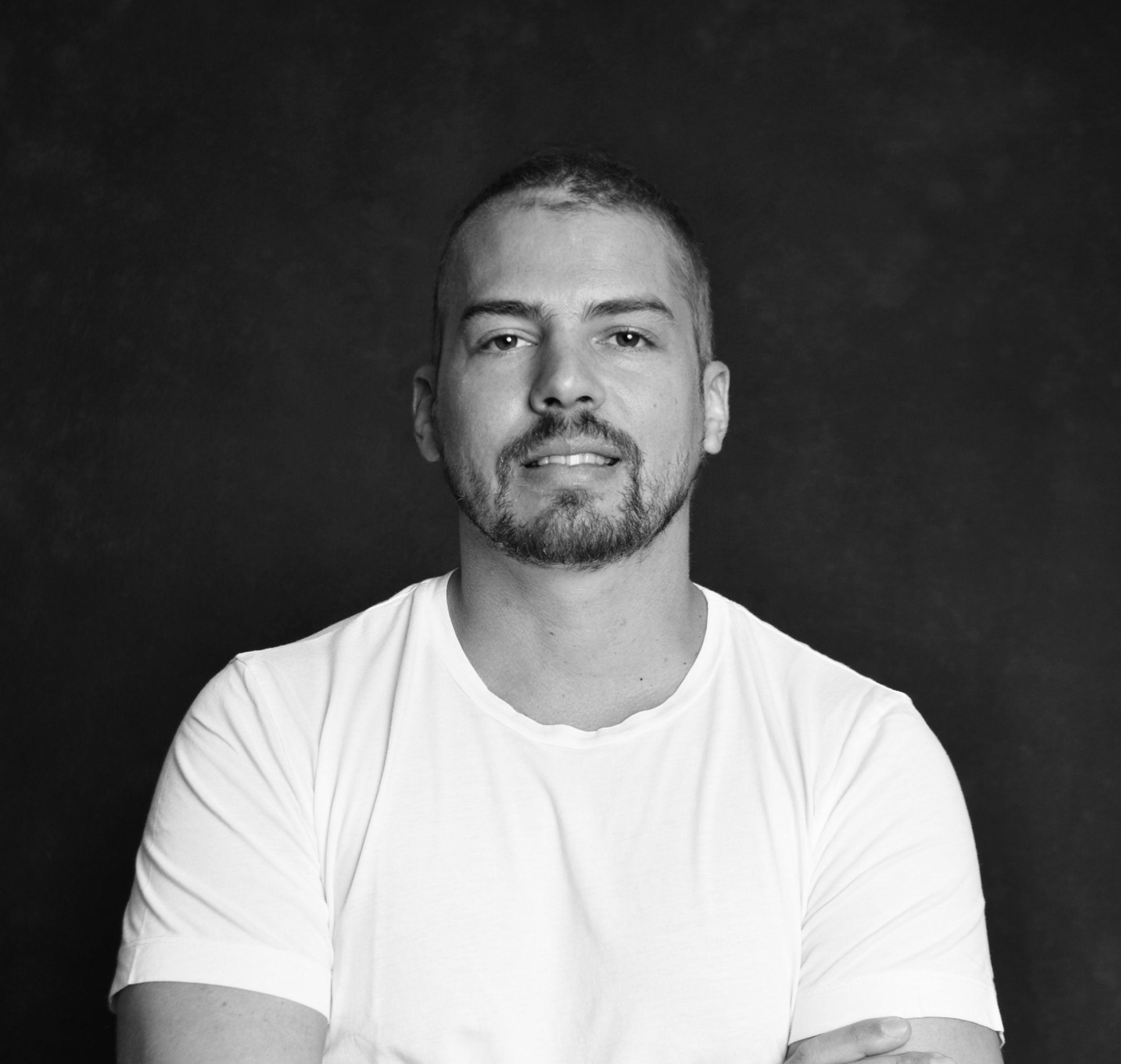 Black and white portrait of a man with short hair and a beard, wearing a white t-shirt, looking at the camera against a dark background.